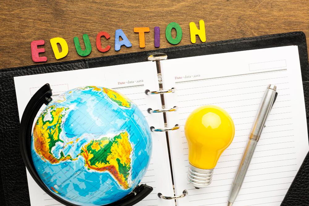 A desk globe, a yellow light bulb and a pen resting on an open notebook, with the word Education spelled in coloured letters above.