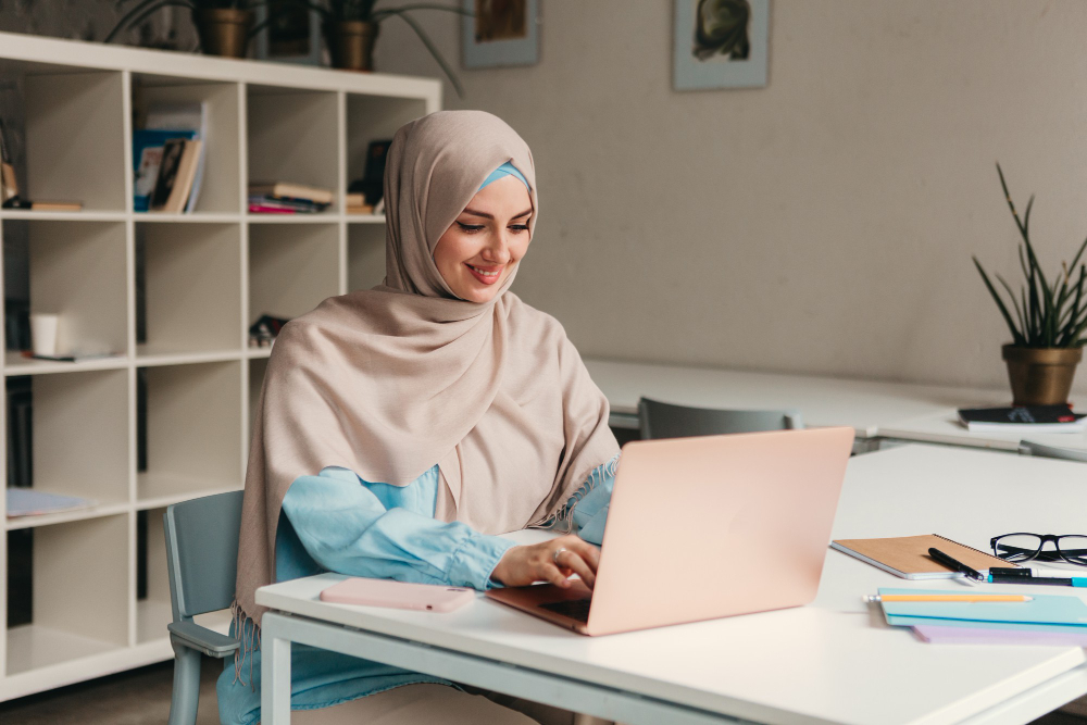 A woman in a hijab smiling as she works at a laptop in a bright study room lined with shelves.