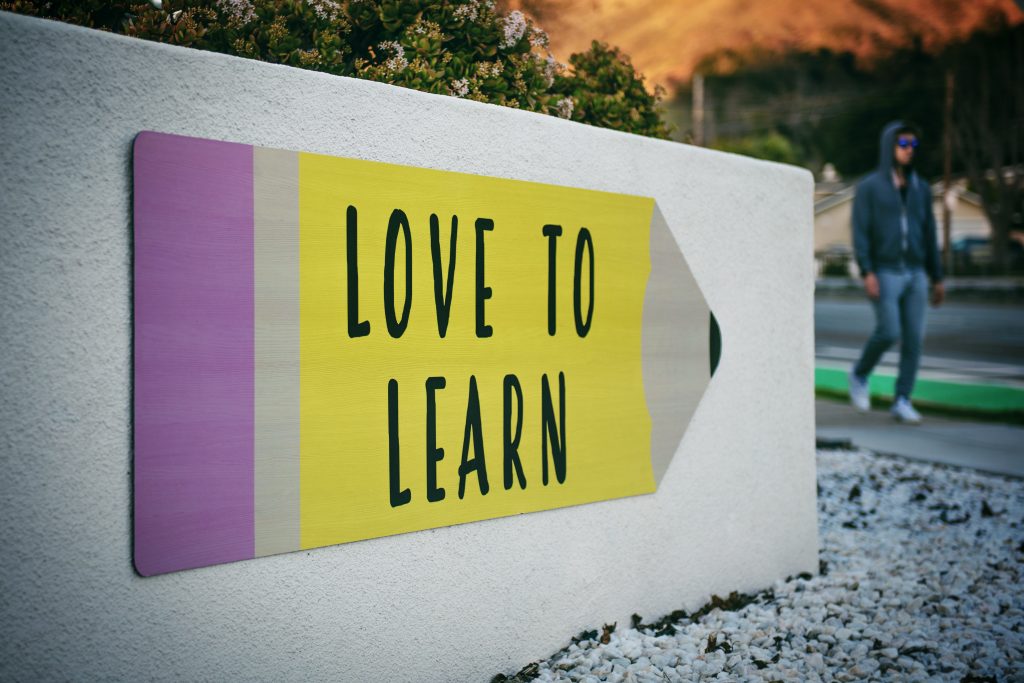 A wall-mounted sign shaped like a yellow pencil reading "Love to learn", with a person walking past at dusk.